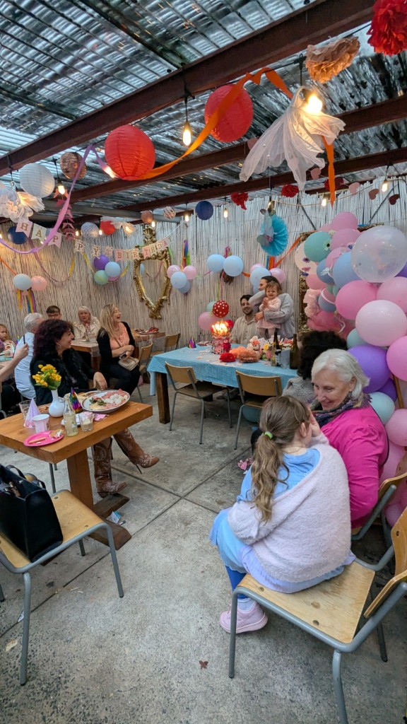 A birthday gathering in the Big Elma courtyard, with lanterns, balloons and a long table.